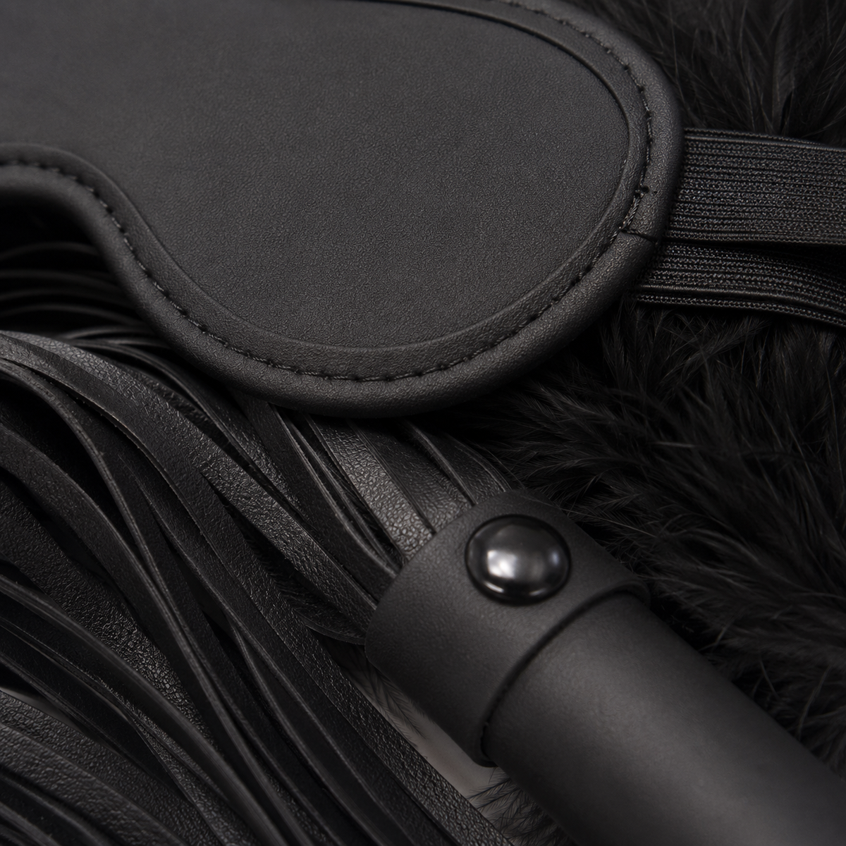 Detailfoto van het zwarte oogmasker, de kunstleren flogger en zachte veren van BondageKit 2